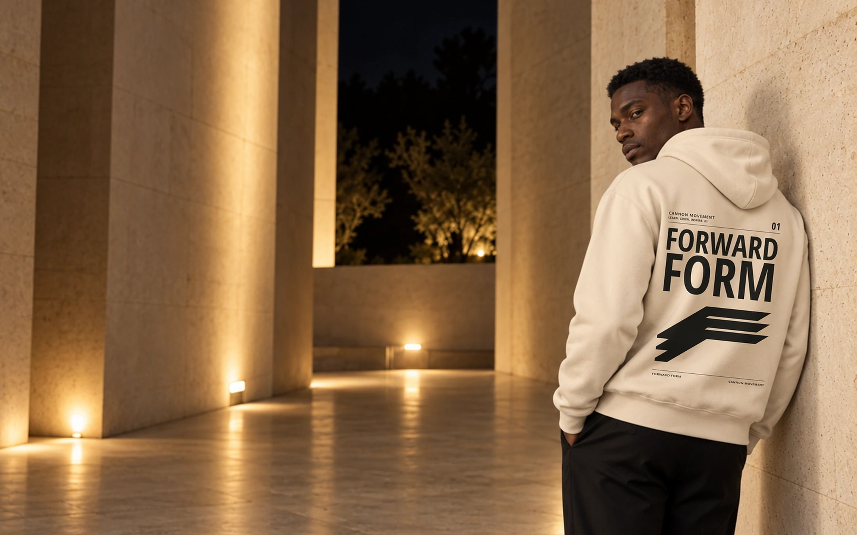 A person in a dust-colored Forward Form hoodie beside illuminated stone architecture at night.